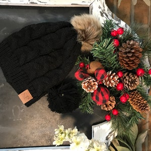 May include: Black knit beanie with a faux fur pom pom and a brown leather patch with the letters "CF" on it. The beanie is sitting on a white painted surface with a Christmas wreath in the background.
