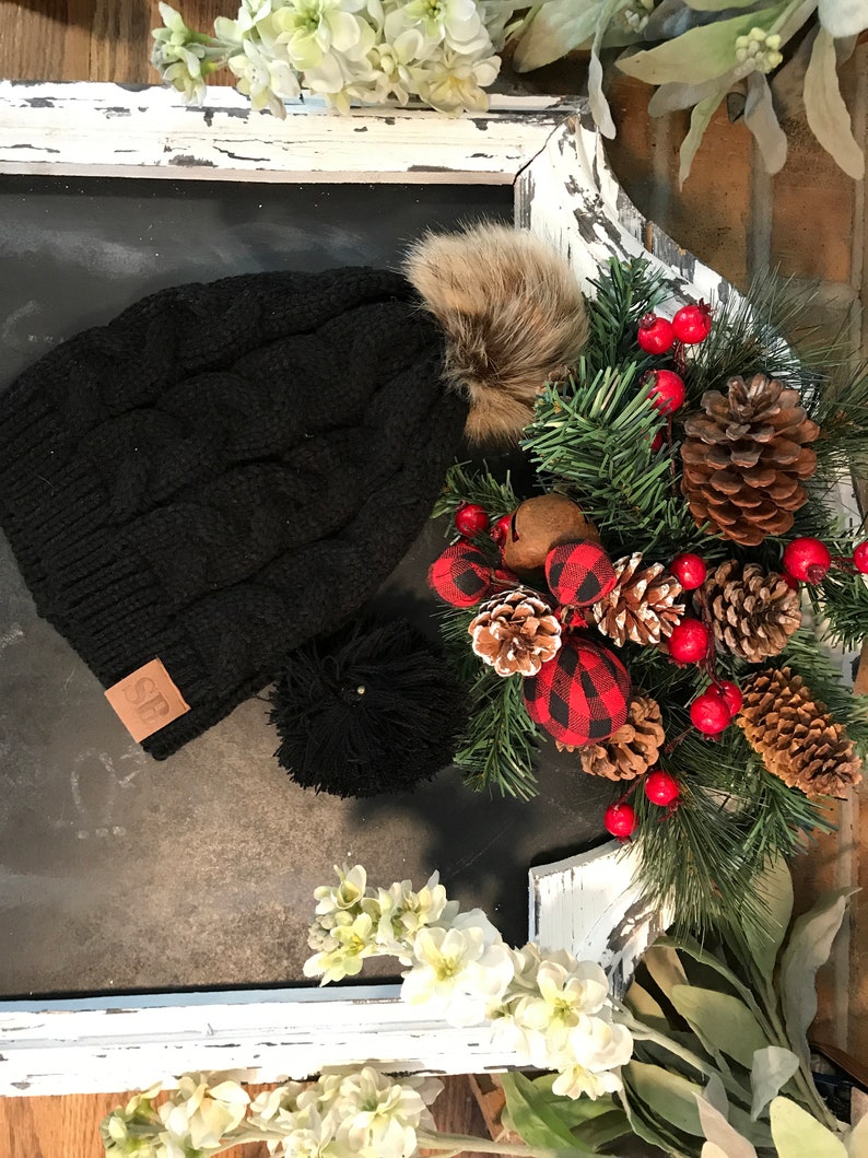 May include: Black knit beanie with a faux fur pom pom and a brown leather patch with the letters "CF" on it. The beanie is sitting on a white painted surface with a Christmas wreath in the background.