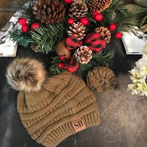 May include: A brown knit beanie with a faux fur pom pom and a brown leather patch with the initials "SB" on it. The beanie is sitting on a black surface with a Christmas wreath in the background.