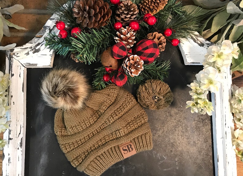 May include: A brown knit beanie with a faux fur pom pom and a brown leather patch with the initials "SB" on it. The beanie is sitting on a black surface with a Christmas wreath in the background.