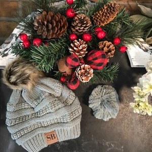 May include: A gray knitted beanie with a brown faux fur pom pom and a brown leather patch with the initials "SB". The beanie is on a black surface with a Christmas-themed arrangement of pine branches, pine cones, red berries, and red and black plaid ornaments.