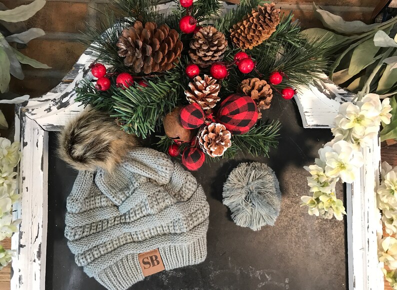May include: A gray knitted beanie with a brown faux fur pom pom and a brown leather patch with the initials "SB". The beanie is on a black surface with a Christmas-themed arrangement of pine branches, pine cones, red berries, and red and black plaid ornaments.