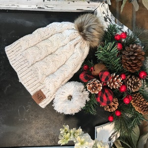 May include: A white cable knit beanie with a faux fur pom pom and a brown leather patch with the initials "SB". The beanie is sitting on a black surface with a white frame and a Christmas-themed garland.