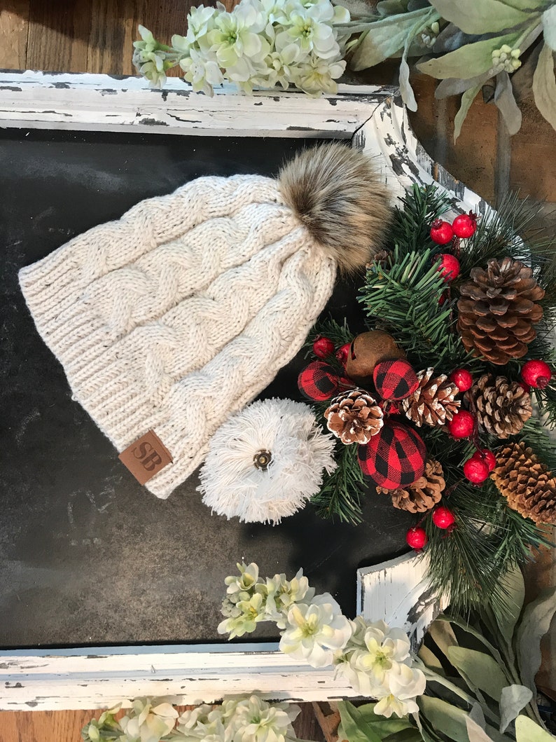 May include: A white cable knit beanie with a faux fur pom pom and a brown leather patch with the initials "SB". The beanie is sitting on a black surface with a white frame and a Christmas-themed garland.