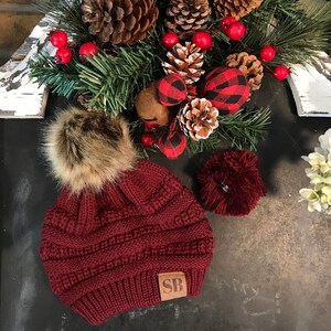 May include: A burgundy knit beanie with a faux fur pom pom and a brown leather patch with the initials "SB".