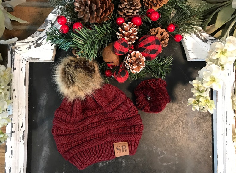 May include: A burgundy knit beanie with a faux fur pom pom and a brown leather patch with the initials "SB".
