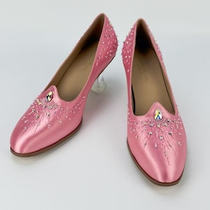 May include: A pair of pink satin court shoes featuring a clear acrylic heel and a pointed toe. The shoes are embellished with intricate silver beadwork and a single iridescent teardrop-shaped crystal centred on the vamp of each shoe.