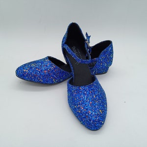 May include: A pair of blue glitter shoes with an ankle strap. The shoes have a pointed toe and a low heel. The brand name "Kay Jane Designs" is visible on the strap.