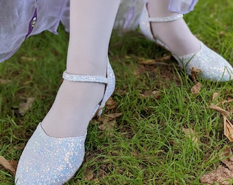 May include: A pair of light blue sparkly shoes with white straps and heels. The shoes are on a bed of green grass.