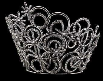 May include: A silver-toned crown featuring an intricate openwork design composed of circular loops and star-shaped accents. The entire structure is encrusted with numerous small, clear, reflective rhinestones that catch the light.