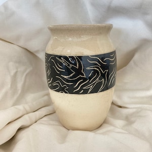 Pode incluir: Um vaso de cerâmica de cor creme com uma faixa preta e um design abstrato. O vaso tem um corpo arredondado e uma abertura ligeiramente alargada.