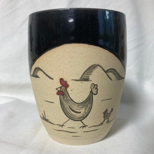 Pode incluir: Uma caneca de cerâmica com um esmalte preto e um desenho creme de uma galinha e um pintinho caminhando em um campo com colinas ao fundo.