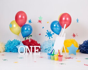 May include: A white wooden easel with a blank canvas, surrounded by colorful balloons, tissue paper pom-poms, and paint splatters. The word "ONE" is spelled out in white letters on the floor.