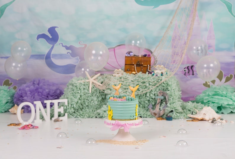 May include: A light blue and pink mermaid-themed cake with a white frosting and two mermaid tails on top. The cake is on a white cake stand with a pink ribbon. The cake is in front of a backdrop with a mermaid, seashells, and a treasure chest.