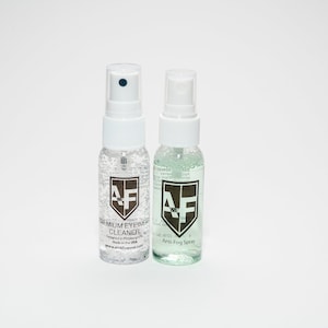 May include: Two small spray bottles with white caps and labels. The bottle on the left is clear and says "Premium Eyewear Cleaner" with a logo of "AF" in a shield. The bottle on the right is green and says "Anti-Fog Spray" with the same logo.