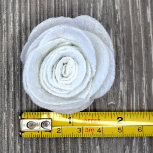 May include: A light blue felt flower, approximately 2 inches in diameter, with layered petals. The flower is positioned on a wooden surface, with a yellow measuring tape alongside it.