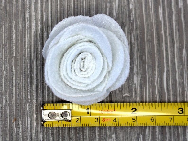 May include: A light blue felt flower, approximately 2 inches in diameter, with layered petals. The flower is positioned on a wooden surface, with a yellow measuring tape alongside it.