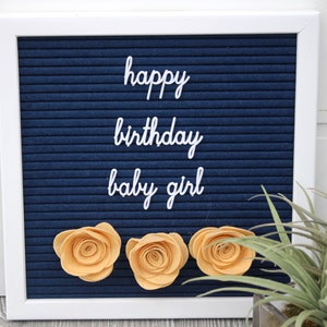 May include: A blue letter board with white letters spelling "happy birthday baby girl". Three yellow felt flowers are attached to the board.