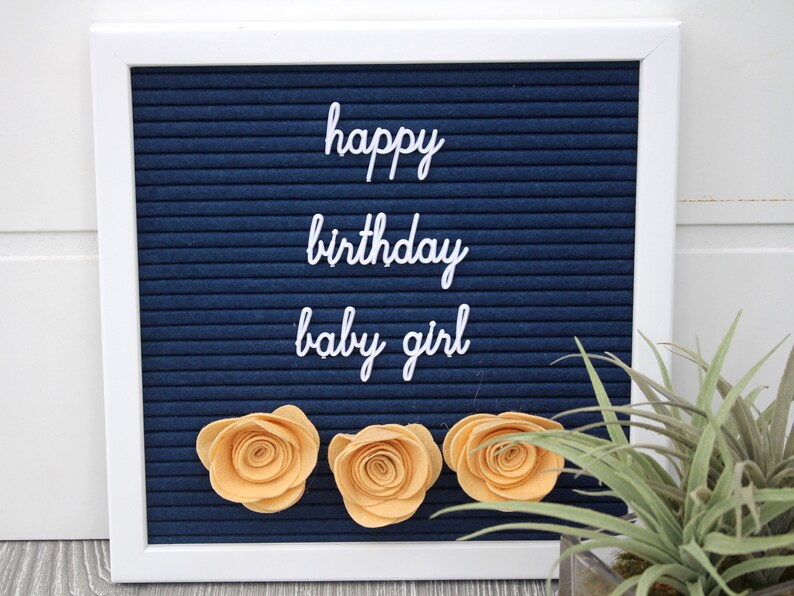 May include: A blue letter board with white letters spelling "happy birthday baby girl". Three yellow felt flowers are attached to the board.