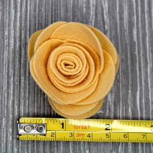 May include: A yellow felt flower with a spiral center. The flower is about 3 inches in diameter and is sitting on a gray wooden surface. A yellow measuring tape is below the flower.
