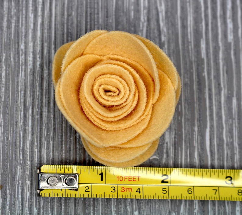May include: A yellow felt flower with a spiral center. The flower is about 3 inches in diameter and is sitting on a gray wooden surface. A yellow measuring tape is below the flower.
