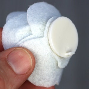 May include: A white, felt-like object with a cream-colored, oval-shaped button attached. The button has a small, protruding tab. The object is held in a person's hand.