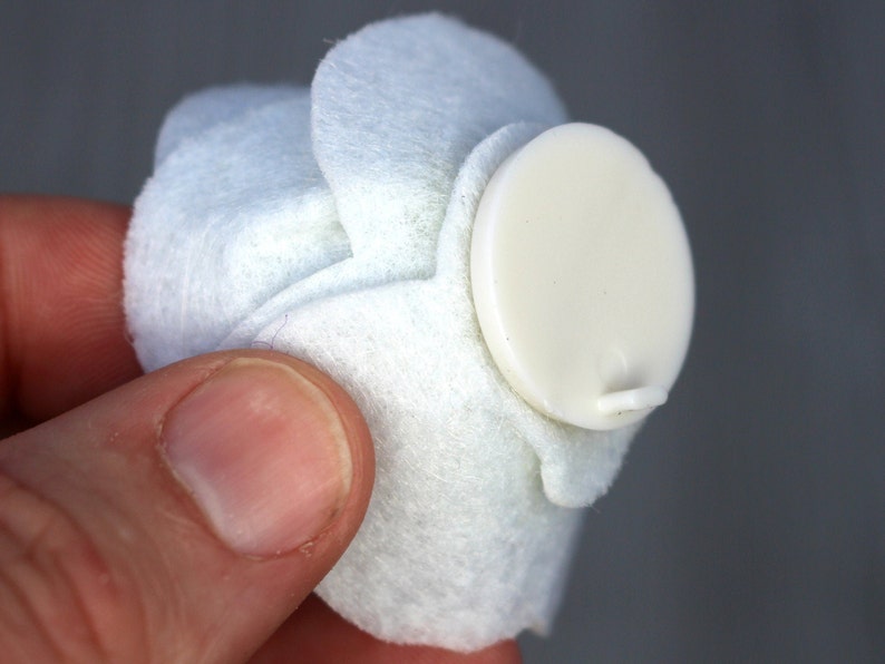 May include: A white, felt-like object with a cream-colored, oval-shaped button attached. The button has a small, protruding tab. The object is held in a person's hand.