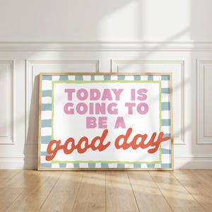 May include: A framed art print with the text "TODAY IS GOING TO BE A good day." The text is in pink and red, set against a white background with a green and blue checkered border. The frame is a light wood color.
