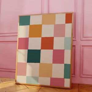 K&ouml;nnte beinhalten: Ein gerahmtes Kunstwerk mit einem Raster aus farbigen Quadraten in T&uuml;rkis, Orange, Rosa und Creme. Das Kunstwerk hat einen goldenen Rahmen und lehnt an einer rosa Wand. Das abstrakte Design erinnert an ein Schachbrettmuster.