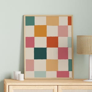 K&ouml;nnte beinhalten: Ein gerahmter abstrakter Kunstdruck mit einem Raster aus farbigen Quadraten in T&uuml;rkis-, Orange-, Rosa- und Cremet&ouml;nen. Das Kunstwerk wird auf einem Holzschrank ausgestellt, mit einer Lampe und zwei wei&szlig;en zylindrischen Objekten im Hintergrund.