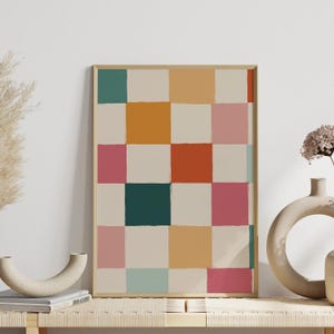 K&ouml;nnte beinhalten: Ein gerahmter Kunstdruck mit einem abstrakten Muster aus farbigen Quadraten. Die Quadrate sind in T&uuml;rkis-, Orange-, Rosa- und Cremet&ouml;nen gehalten, mit einem beigen Hintergrund. Das Kunstwerk wird auf einer Holzbank mit dekorativen Vasen ausgestellt.
