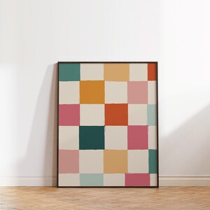 K&ouml;nnte beinhalten: Ein gerahmtes Kunstwerk mit einem farbenfrohen Schachbrettmuster. Die Quadrate sind in verschiedenen Farben wie T&uuml;rkis, Orange, Rosa und Creme bemalt, mit sichtbaren Pinselstrichen. Das Kunstwerk lehnt an einer wei&szlig;en Wand auf einem Holzboden.