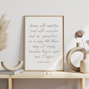 May include: A framed print with a gold border featuring a quote by Kamala Harris in black script: "dream with ambition, lead with conviction..." The print is displayed on a light-colored wooden table with decorative vases and dried flowers.