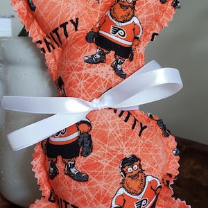 Puede incluir: Un conejito de tela naranja hecho a mano con un lazo de cinta blanca. El conejito está decorado con un patrón repetido de la mascota de los Flyers de Filadelfia, Gritty, y la palabra "Gritty".