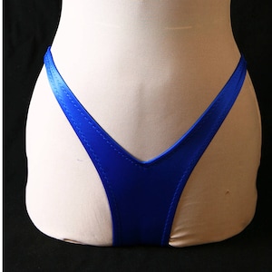 Puede incluir: Braguita de bikini azul real con forma de V en la parte delantera y trasera. La tela tiene un ligero brillo y los bordes están cuidadosamente cosidos. La prenda se muestra en un maniquí sobre un fondo negro.