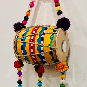 May include: A decorative wall hanging featuring a yellow drum with blue, red, and white painted stripes, accented by gold strings. Three long strands of colorful beads, small circular mirrors, and brass bells hang from the bottom of the drum.