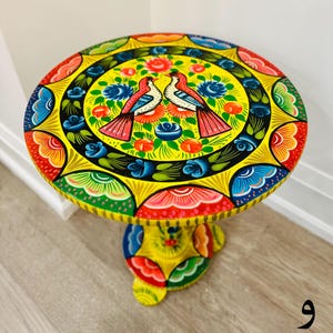 May include: A yellow painted wooden table with a round top and a pedestal base. The top is decorated with a colorful floral design and two birds. The base is also decorated with a colorful floral design.