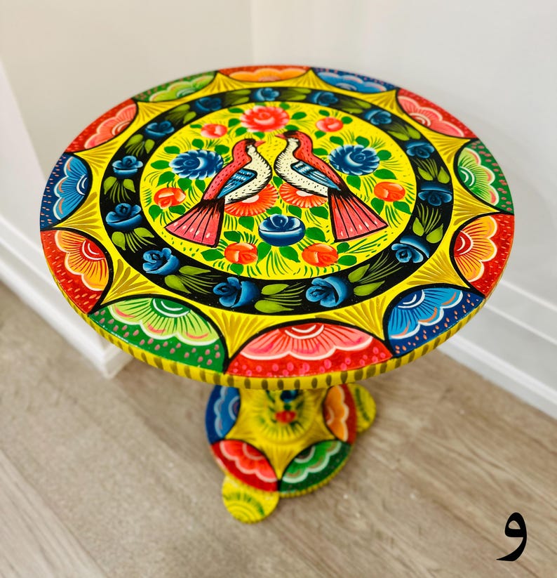 May include: A yellow painted wooden table with a round top and a pedestal base. The top is decorated with a colorful floral design and two birds. The base is also decorated with a colorful floral design.