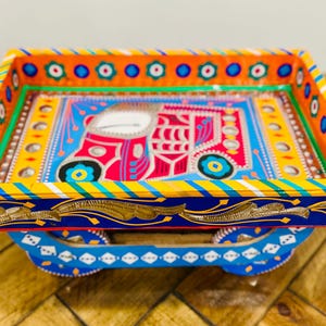 May include: A rectangular wooden tray featuring vibrant hand-painted designs in orange, blue, red, and yellow. The interior base displays a stylised vehicle illustration, while the exterior sides are decorated with gold floral patterns.