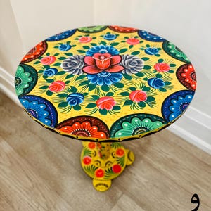 Puede incluir: Una mesa redonda amarilla con un diseño floral pintado a mano. La superficie de la mesa presenta un motivo floral central con rosas coloridas, hojas y espirales. La base de la mesa también está pintada con un diseño floral.