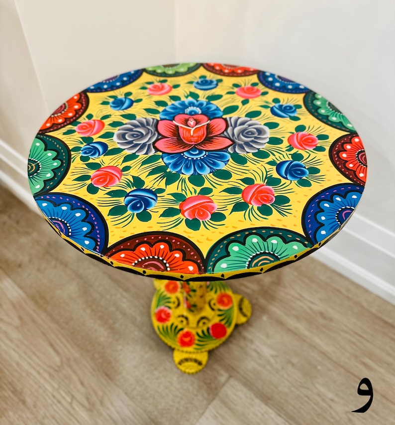 May include: A yellow round table with a hand-painted floral design. The table top features a central floral motif with colorful roses, leaves, and swirls. The base of the table is also painted with a floral design.