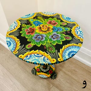 May include: A hand-painted side table with a black base and a round top. The top is decorated with a colorful floral design featuring yellow, red, blue, and green flowers. The table has a unique, traditional style.