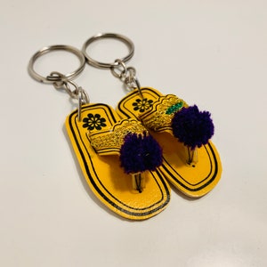 May include: A pair of yellow leather flip-flop keychains with purple and green pom-poms. The keychains are attached to silver key rings.
