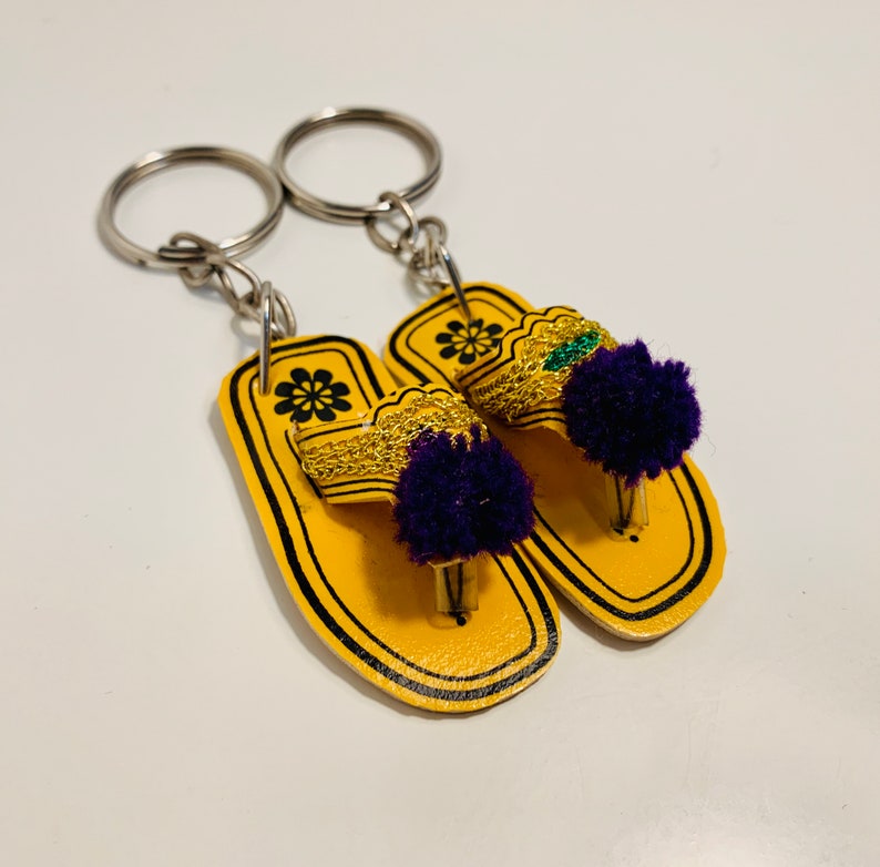 May include: A pair of yellow leather flip-flop keychains with purple and green pom-poms. The keychains are attached to silver key rings.