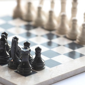 May include: A chess set featuring polished black and white stone pieces arranged on a marble chessboard with alternating black and white squares. The pieces include pawns, rooks, knights, bishops, queens, and kings, all set in a starting position.