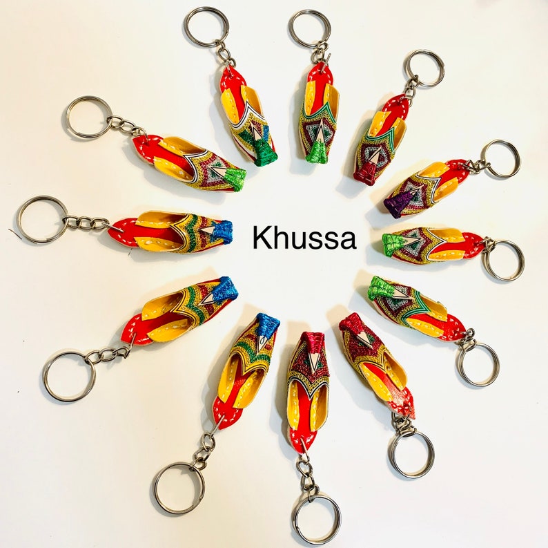 May include: A set of 12 colorful keychains featuring miniature embroidered Khussa shoes. The shoes are decorated with intricate patterns and have a red, yellow, green, and blue color scheme. The word "Khussa" is written in the center of the image.