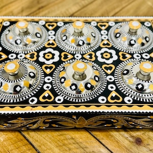 May include: A decorative wooden serving tray with six lidded compartments. The tray is black with gold and white accents, featuring floral and geometric patterns. The lids are silver with gold knobs.