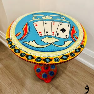 May include: A hand-painted wooden side table with a blue, yellow, and red design. The table top features four playing cards, all aces, with a red heart, a black spade, and two red diamonds. The table base is red with a floral design.