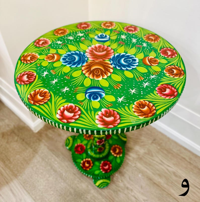 May include: A green painted side table with a floral design. The table top features a circular design of flowers in shades of red, blue, and yellow. The base of the table is also painted green with a floral design.
