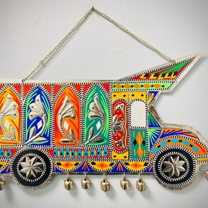 May include: A decorative metal wall hanging shaped like a truck, featuring vibrant hand-painted patterns in orange, blue, green, and yellow. The piece includes embossed floral motifs, two circular wheels, and small metal bells hanging from the bottom.